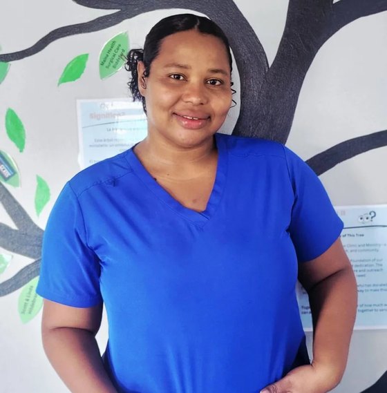 Nurse-intern Kilcia, wearing blue scrubs, in front of the clinic's tree mural.